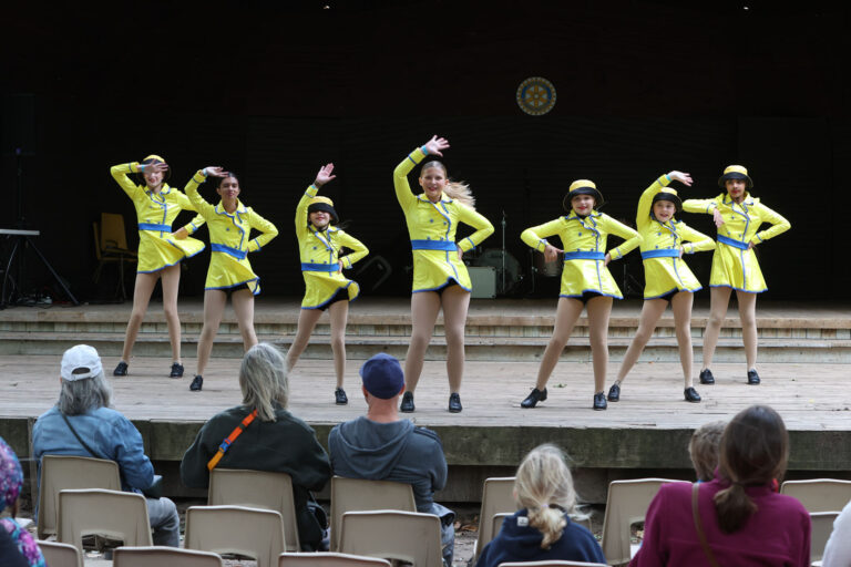 2024 Uxbridge Fair - Seven performers in bright yellow dance costumes with blue trim strike coordinated pose on wooden stage at Uxbridge Fair, with seated audience members watching from plastic chairs in the foreground.