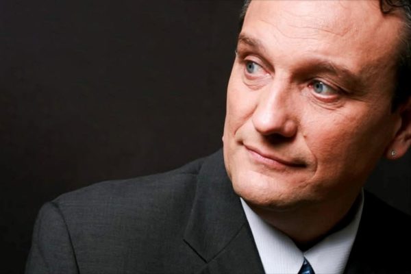 Professional headshot of Robert Maxwell, comedy hypnotist, wearing a dark business suit with a white shirt and blue tie, smiling warmly at the camera