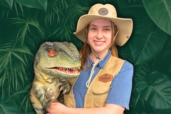 Smiling female park ranger in tan vest and wide-brimmed hat holding a realistic Velociraptor puppet with orange eyes and detailed green scales against a lush tropical plant background.