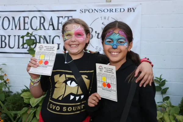 Two children with festival face paint smiling and holding Uxbridge Fair scavenger hunt cards in front of the Homecraft Building.