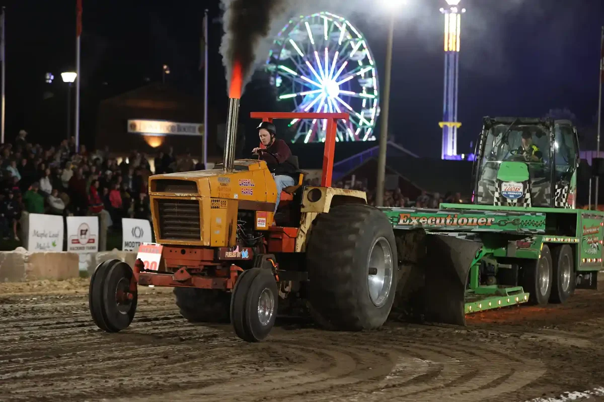 Tractor pulling a weighted sled at the Uxbridge Fair Tractor Pull, midway Ferris wheel lit up behind