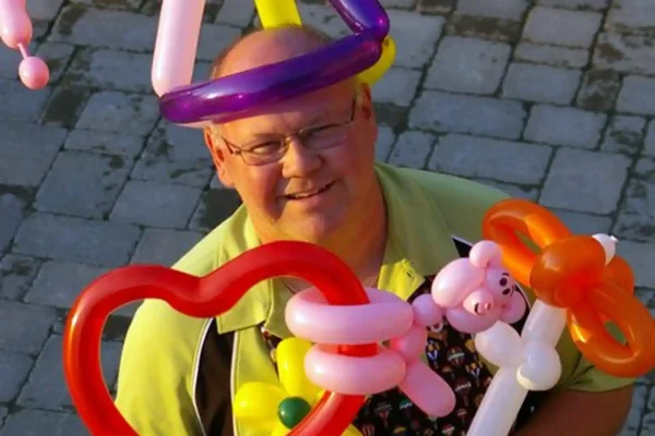 Balloon twister Steve Fisher smiling while wearing a purple and yellow balloon crown, surrounded by colourful balloon animal creations including a heart, a teddy bear, and a butterfly